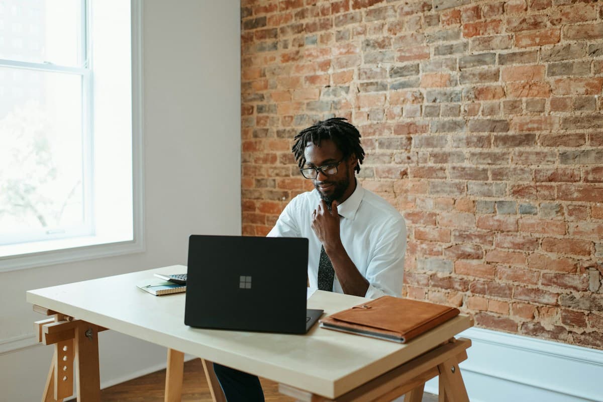 A dark-skinned man managing work at a laptop in a sunlit workspace